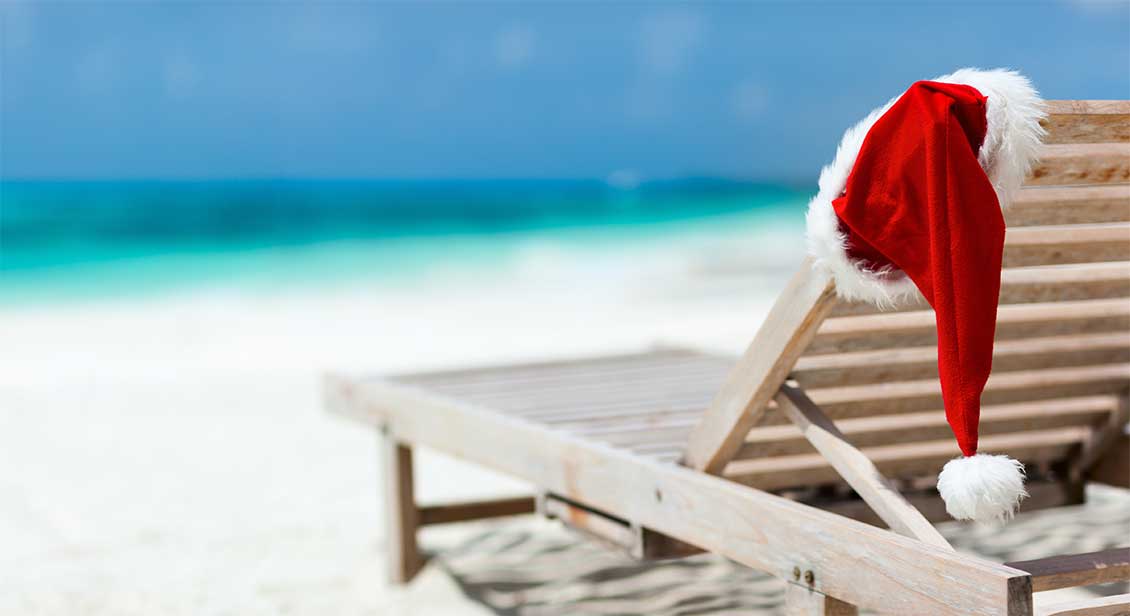 A Santa hat draped on a wooden lounger situated on a beach with the sea in the background.